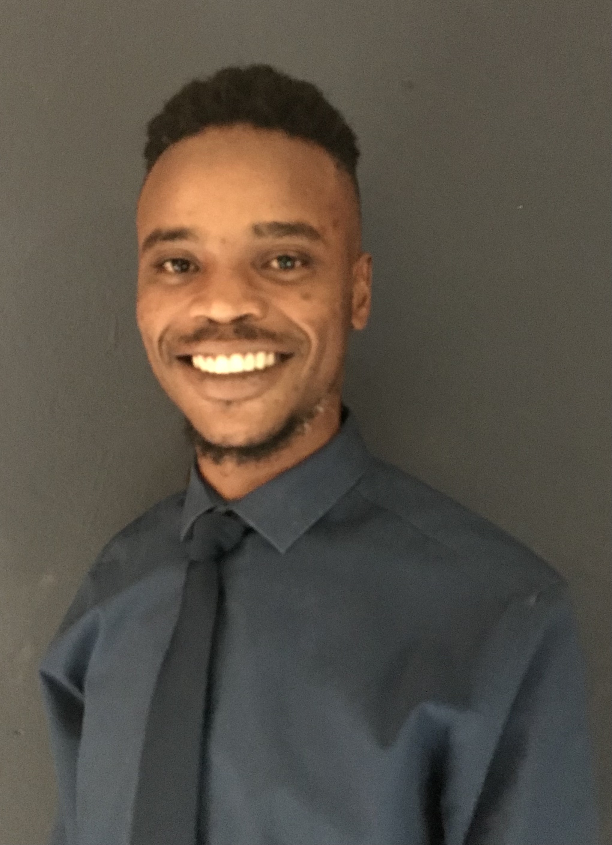 Mbhoni wearing a purple shirt and a black tie, smiling for a professional photoshoot.